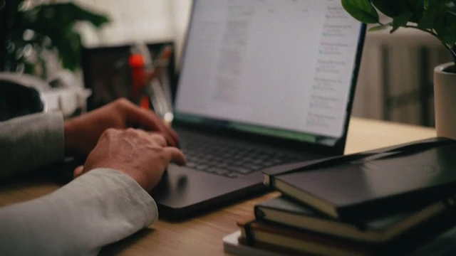 Hands typing on a laptop beside a stack of books at Jon’s home